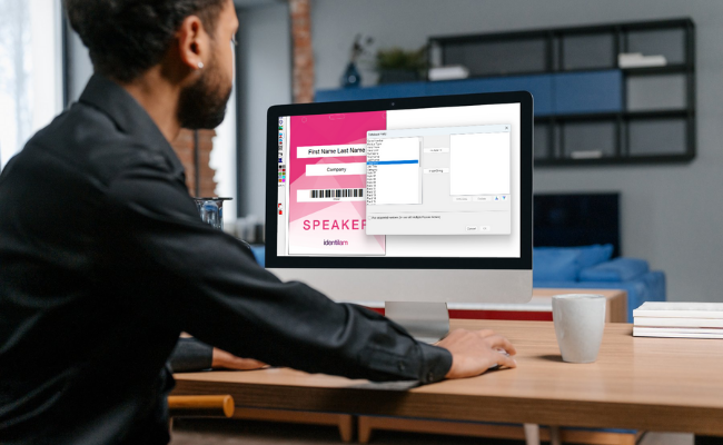 A man designing a custom name badge on a desktop computer using badge creation software, with a &ldquo;Speaker&rdquo; template visible on screen