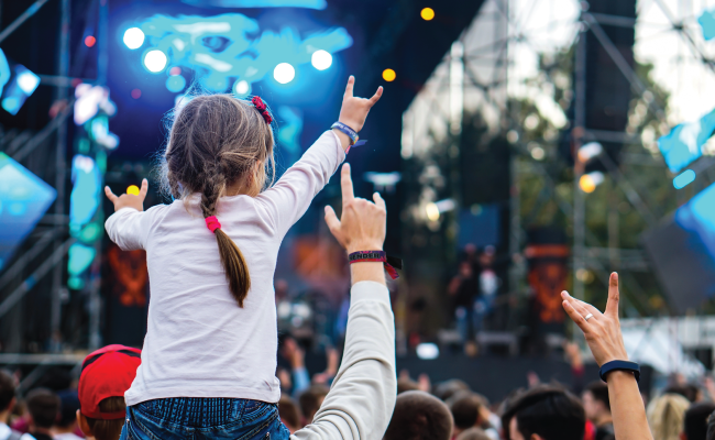 Young girl wearing a safety wristband sitting on adult's shoulders at a music festival.